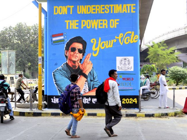 People walk past a pillar with a portrait of Bollywood actor Shah Rukh Khan and slogans to encourage people to cast their vote for the Lok Sabha election (File Photo/ANI)