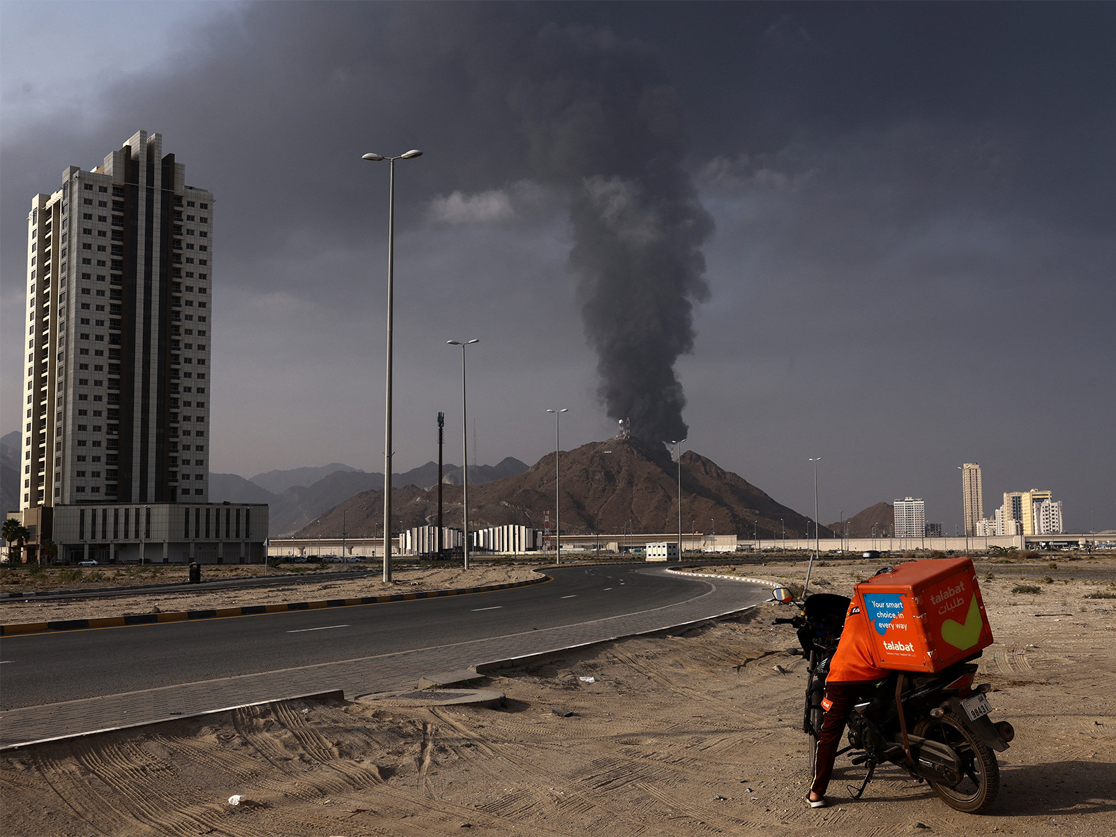 A man stands beside a motorcycle as smoke rises from the Fujairah Oil Industry Zone after debris from an intercepted drone caused a fire, amid the US-Israel conflict with Iran, in Fujairah, UAE. (Photo/Reuters)